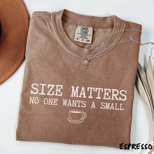 May include: Espresso-colored t-shirt with the text "SIZE MATTERS NO ONE WANTS A SMALL" and a coffee cup graphic. A silver necklace with a pendant is draped across the shirt. The shirt is a Comfort Colors brand.