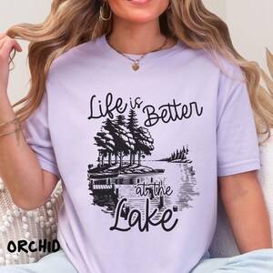 Puede incluir: Una camiseta lavanda con un estampado gr&aacute;fico negro que dice "Life is Better at the Lake". El gr&aacute;fico presenta una escena de lago con &aacute;rboles y un muelle.