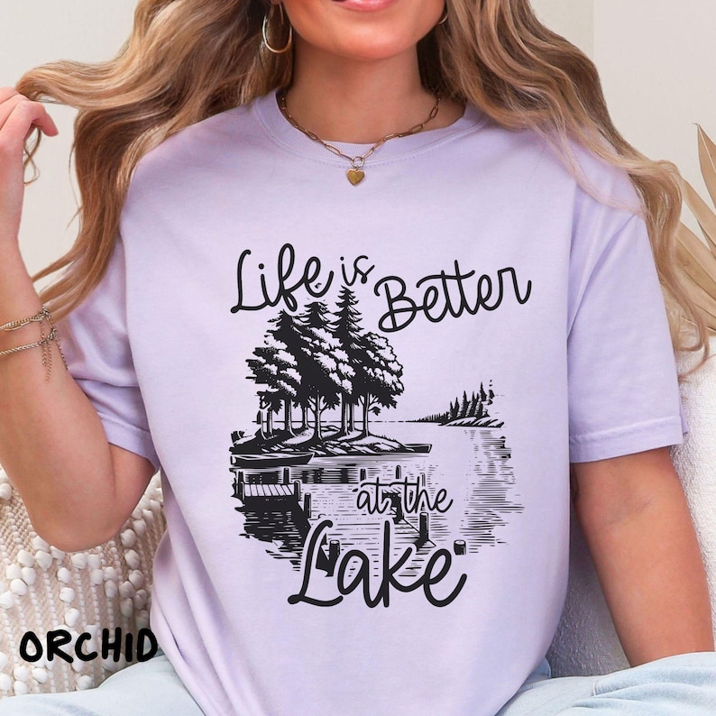 Puede incluir: Una camiseta lavanda con un estampado gr&aacute;fico negro que dice "Life is Better at the Lake". El gr&aacute;fico presenta una escena de lago con &aacute;rboles y un muelle.