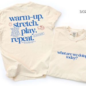 May include: Two ivory-colored t-shirts. One shirt has the text "warm-up, stretch, play, repeat" in blue, with additional text and graphics. The other shirt has the text "what are we doing today?" in blue.