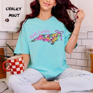 May include: A turquoise t-shirt with the text "North Carolina" and a floral design in the shape of the state. The shirt also features a pink bow. The words "Chalky Mint" are printed in the upper left corner.