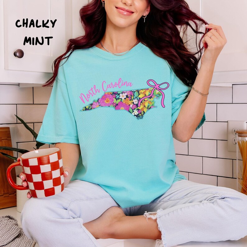 May include: A turquoise t-shirt with the text "North Carolina" and a floral design in the shape of the state. The shirt also features a pink bow. The words "Chalky Mint" are printed in the upper left corner.