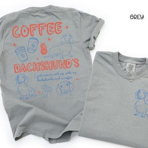 May include: Two grey t-shirts. One displays the text "COFFEE & DACHSHUND'S" with dachshund and coffee cup illustrations. The other shirt has a dachshund illustration. Both shirts have blue and red designs.