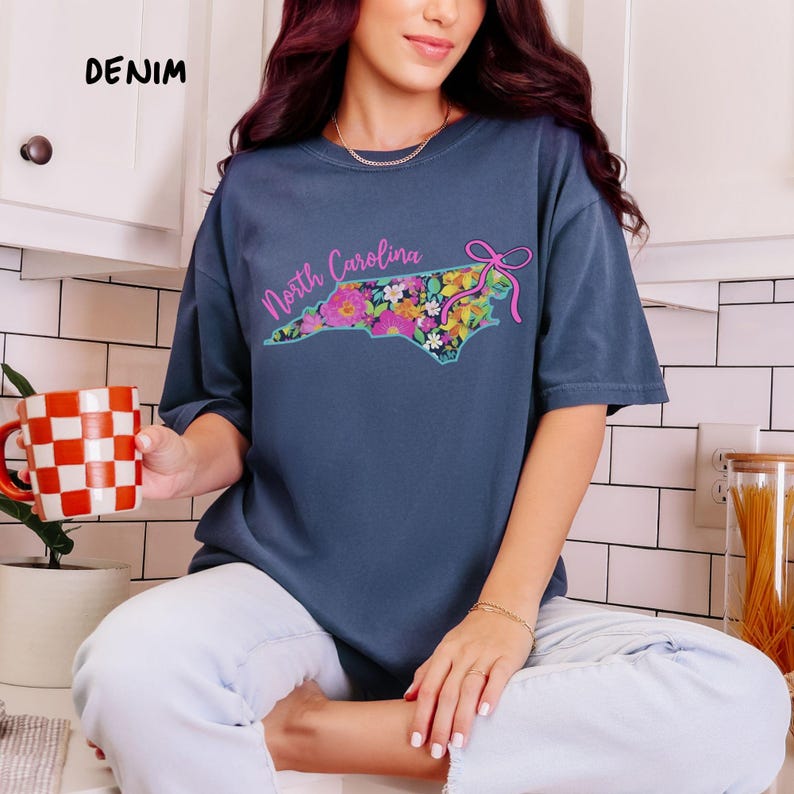 May include: Denim blue t-shirt featuring a floral North Carolina state outline design. The state outline is filled with colorful flowers and a pink bow. The text "North Carolina" is above the design.