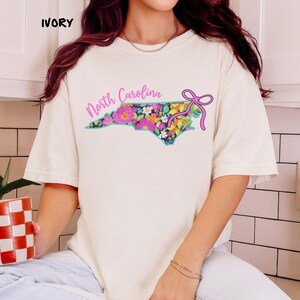 May include: Ivory-colored t-shirt featuring a floral North Carolina state outline design. The state outline is filled with colorful flowers and a teal border. The words "North Carolina" are written in pink script above the design, with a pink bow on the right.