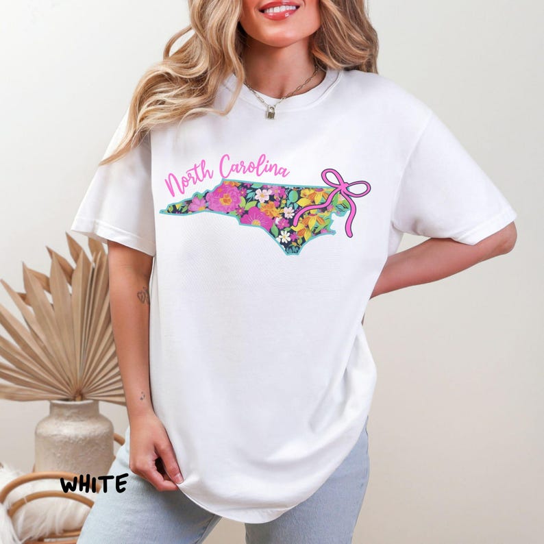 May include: White t-shirt featuring a floral North Carolina state outline with the text "North Carolina" in pink. A pink bow is on the right side of the state outline. The word "WHITE" is at the bottom left of the shirt.