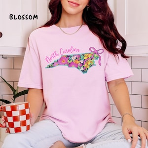 May include: A light pink t-shirt with a floral North Carolina state outline design. The design includes the words "North Carolina" in a script font and a pink bow. The word "BLOSSOM" is printed in black at the top left.