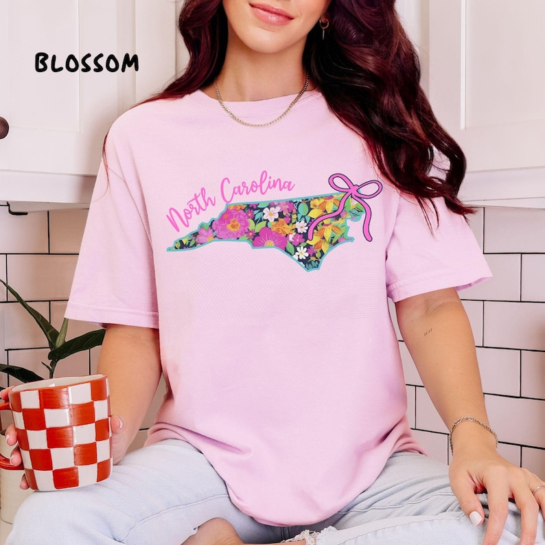 May include: A light pink t-shirt with a floral North Carolina state outline design. The design includes the words "North Carolina" in a script font and a pink bow. The word "BLOSSOM" is printed in black at the top left.