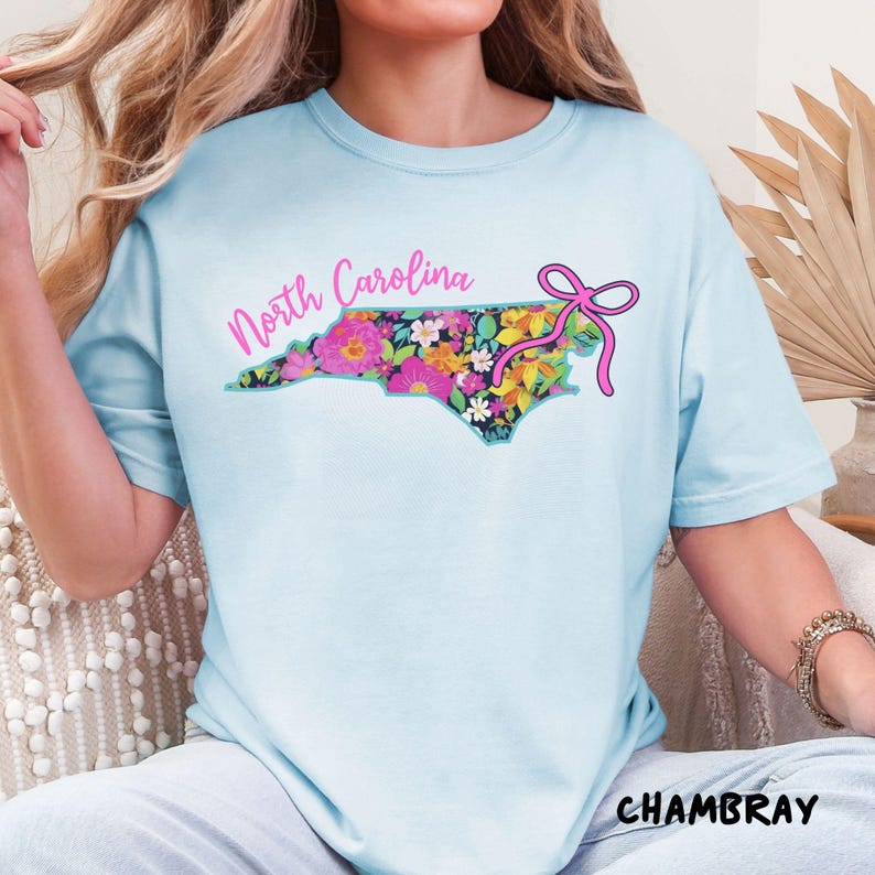 May include: Light blue t-shirt with a floral North Carolina state outline design. The design features a variety of colorful flowers and a pink bow. The word "North Carolina" is written in pink script above the state outline. The word "CHAMBRAY" is written in black block letters below.
