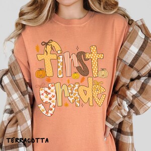 Op de afbeelding: Terracotta-kleurig T-shirt met de woorden "First Grade" in een speels, herfstthema-ontwerp. De tekst bevat pompoenen, bladeren en verschillende patronen in tinten oranje, bruin en geel. Het shirt heeft een ronde hals.