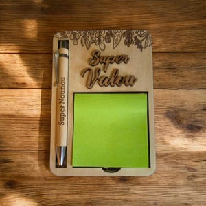 Peut inclure: Un ensemble bloc-notes et stylo en bois. Le bloc-notes contient un bloc de notes adhésives vert et les mots "Super Valou" et "Super Nounou" gravés. Le stylo est en bambou avec une pointe argentée.