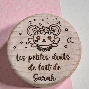 Peut inclure: Un couvercle rond en bois clair avec une gravure laser représentant une souris stylisée portant une couronne de fleurs, entourée d'étoiles et de lunes. Le texte "les petites dents de lait de Sarah" est gravé en écriture cursive sous la souris.