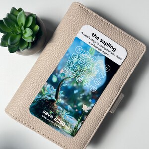 Puede incluir: Una cartera de cuero beige con una tarjeta con un gr&aacute;fico de &aacute;rbol y el texto "the sapling". La tarjeta tiene una cuadr&iacute;cula de n&uacute;meros y el texto "save &pound;250". Una peque&ntilde;a planta suculenta en maceta est&aacute; en el fondo.