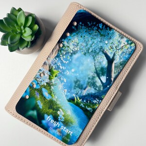 Puede incluir: Una agenda beige claro con una inserci&oacute;n con tem&aacute;tica de la naturaleza que presenta un paisaje azul y verde con flores blancas y el texto "fresh start the cash cove". Una peque&ntilde;a planta suculenta en una maceta gris est&aacute; en la esquina superior izquierda.