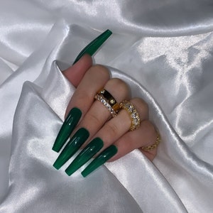 Op de afbeelding: Close-up van een hand met lange, donkergroene, amandelvormige nagels. De hand draagt ​​meerdere gouden ringen met heldere stenen.