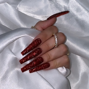 May include: Close-up of long, red glitter nails with a gold ring on the middle finger.