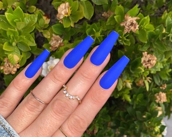 May include: Close-up of a hand with long, blue, matte-finish, coffin-shaped acrylic nails. The nails are all the same length and shape. The hand is wearing a thin gold ring with small diamonds.
