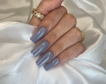 May include: Close-up of a hand with long, square-shaped nails painted a light gray color. The nails are polished to a high shine. The hand is wearing a gold ring with clear stones.