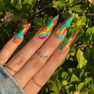 Pode incluir: Close-up de uma mão com unhas longas e pontiagudas pintadas com um design abstrato colorido. As unhas são pintadas em tons de azul, verde, laranja, amarelo e rosa. A mão está usando um anel de ouro.