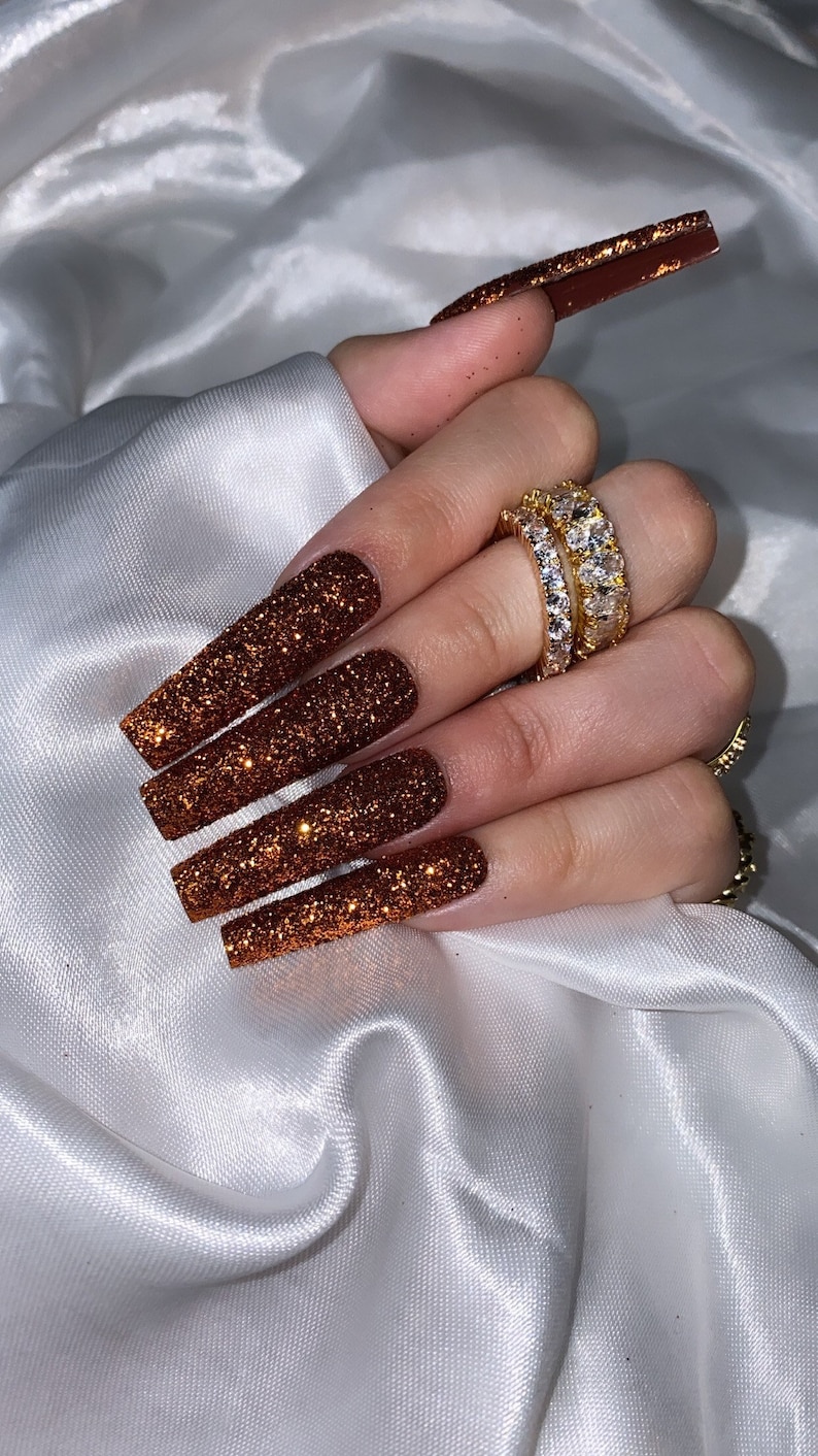 May include: Long, square-shaped, brown glitter nails with a gold glitter accent on the index finger.