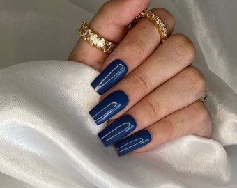 May include: Close-up of hands with long, square-shaped nails painted a deep blue color. The nails are manicured and have a glossy finish. The person is wearing a gold ring with clear stones.