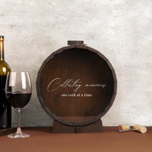 May include: A wooden wine cork holder shaped like a barrel with the text "Collecting memories, one cork at a time."  The barrel is brown and has a metal band around the top. A wine bottle and a wine glass are in the background.