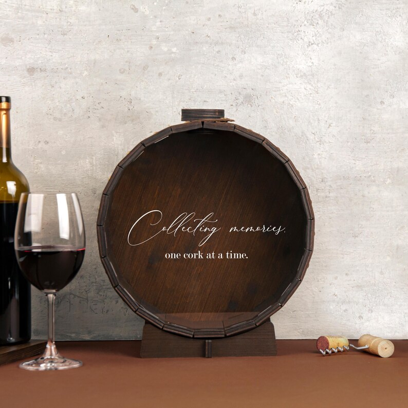 May include: A wooden wine cork holder shaped like a barrel with the text "Collecting memories, one cork at a time."  The barrel is brown and has a metal band around the top. A wine bottle and a wine glass are in the background.