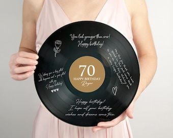 Op de afbeelding: Een zwarte vinylplaat met een gouden middelpunt, gepersonaliseerd met het nummer 70 en de naam "Roger". De plaat is voorzien van verjaardagswensen en decoratieve elementen. De plaat wordt vastgehouden door een persoon in een roze jurk.