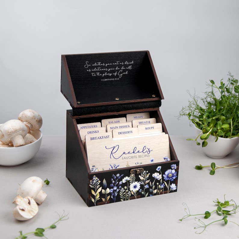May include: A black wooden recipe box with a floral design and a quote from 1 Corinthians 10:31. The box has dividers for different recipe categories, including appetizers, drinks, breakfast, salads, main dishes, desserts, meats, bread & soups. The box is labeled "Rachel's Favorite Recipes".