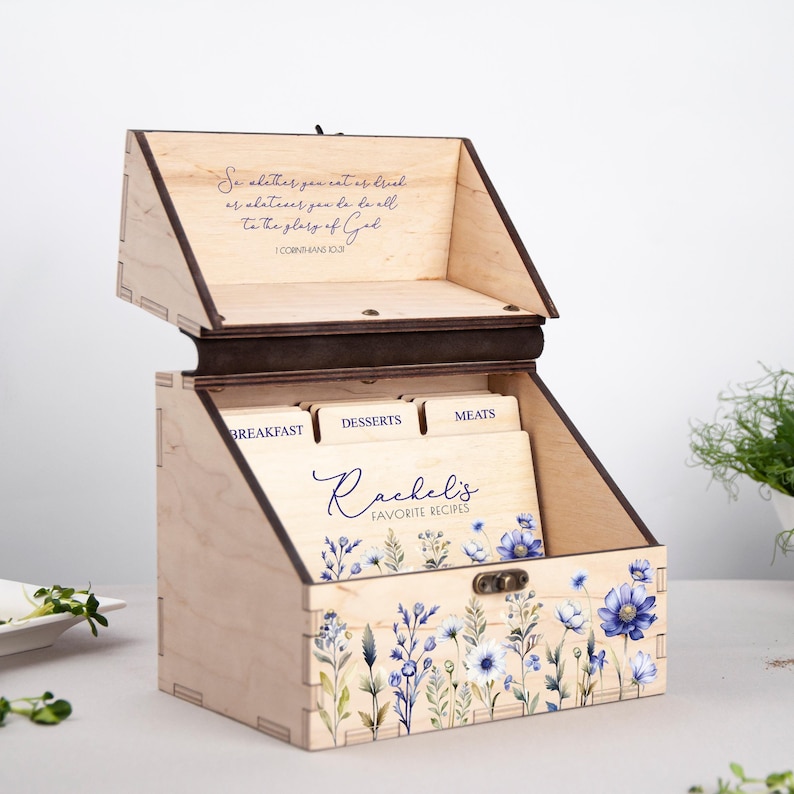 May include: A wooden recipe box with a floral design and the words "Rachel's Favorite Recipes" on the front. The box has four compartments labeled "Breakfast", "Desserts", "Meats", and "Sides". The inside of the box is lined with a brown fabric. The lid of the box has a quote from the Bible: "So whether you eat or drink or whatever you do, do it all to the glory of God." 1 Corinthians 10:31