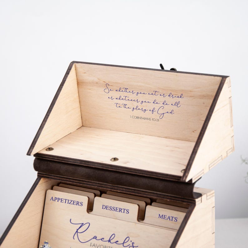 May include: A wooden recipe box with a brown stain and a light wood interior. The box has dividers for different categories of recipes, including appetizers, desserts, and meats. The box is engraved with the text "So whether you eat or drink or whatever you do, do it all to the glory of God" and "1 Corinthians 10:31". The box is also personalized with the name "Rachel's Favorite Recipes".