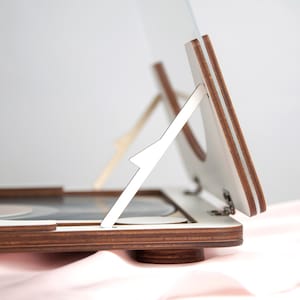 May include: A wooden stand with a white hinged lid and a white adjustable support. The stand is designed to hold a record player.