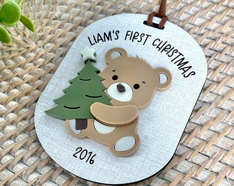 May include: White wooden ornament with a brown teddy bear holding a green Christmas tree. The ornament says "Liam's First Christmas 2016".