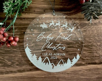 Puede incluir: Adorno de Navidad redondo y transparente con un diseño blanco de un trineo tirado por renos, copos de nieve y árboles. El texto "Baby's First Christmas" está escrito en cursiva, seguido de "Lucas Adrian 2022".