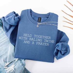 May include: A denim blue crew neck sweatshirt with the text "HELD TOGETHER WITH BALING TWINE AND A PRAYER" in white. A silver necklace with a small pendant is draped across the sweatshirt. Light blue jeans are visible.