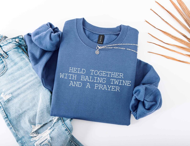 May include: A denim blue crew neck sweatshirt with the text "HELD TOGETHER WITH BALING TWINE AND A PRAYER" in white. A silver necklace with a small pendant is draped across the sweatshirt. Light blue jeans are visible.