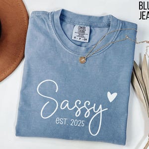 May include: A blue jean coloured t-shirt with the text "Sassy est. 2025" and a small white heart.