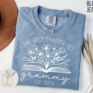 May include: A blue jean-colored t-shirt with the text "The Best Chapter Yet grammy EST. 2026" in white. The design includes an open book with flowers. The shirt has a crew neck and short sleeves.