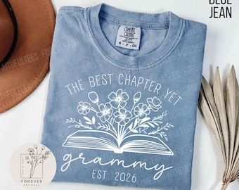 Pode incluir: Uma camiseta azul jeans com o texto branco "The Best Chapter Yet grammy EST. 2026". O design inclui um livro aberto com flores. A camisa tem gola redonda e mangas curtas.