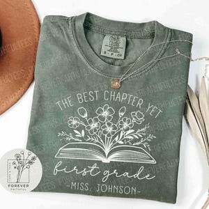 May include: Sage green t-shirt with the text "The Best Chapter Yet" and "first grade - Miss. Johnson" in white script. A floral design is above the words "first grade". A silver necklace with a pendant is on the shirt.