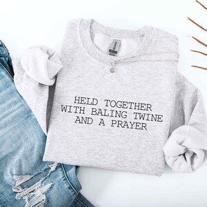 May include: A pale grey sweatshirt featuring the text "HELD TOGETHER WITH BALING TWINE AND A PRAYER" in black. The sweatshirt is styled with light blue denim jeans with distressed details and a silver necklace.