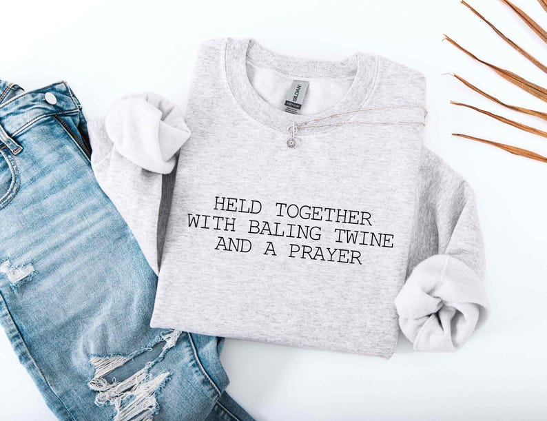 May include: A pale grey sweatshirt featuring the text "HELD TOGETHER WITH BALING TWINE AND A PRAYER" in black. The sweatshirt is styled with light blue denim jeans with distressed details and a silver necklace.