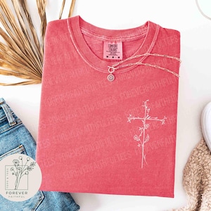 Könnte beinhalten: Korallenfarbenes T-Shirt mit weißem Blumenkreuz-Design und dem Schriftzug "FOREVER FAITHFUL". Eine silberne Halskette mit rundem Anhänger liegt auf dem T-Shirt. Das T-Shirt liegt gefaltet auf einer Jeans. Das Markenlogo ist sichtbar.