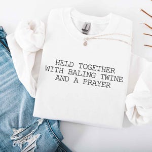May include: A white sweatshirt with the text "HELD TOGETHER WITH BALING TWINE AND A PRAYER" in black. The sweatshirt is styled with blue jeans and a delicate necklace. The image has a casual, lifestyle aesthetic.