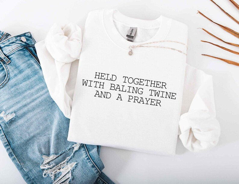 May include: A white sweatshirt with the text "HELD TOGETHER WITH BALING TWINE AND A PRAYER" in black. The sweatshirt is styled with blue jeans and a delicate necklace. The image has a casual, lifestyle aesthetic.