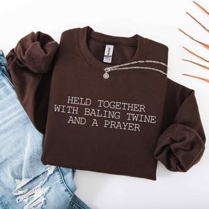 May include: A dark brown sweatshirt with the text "HELD TOGETHER WITH BALING TWINE AND A PRAYER" in white. A silver necklace with a pendant is draped across the sweatshirt. Light blue jeans are visible in the image.