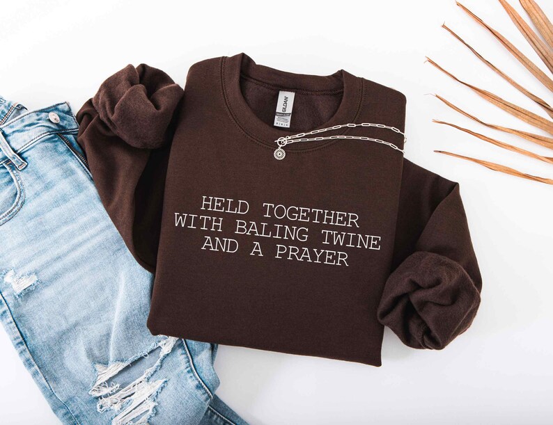 May include: A dark brown sweatshirt with the text "HELD TOGETHER WITH BALING TWINE AND A PRAYER" in white. A silver necklace with a pendant is draped across the sweatshirt. Light blue jeans are visible in the image.