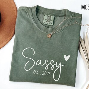 May include: A moss green t-shirt with the text "Sassy est. 2025" and a small white heart printed on the front.