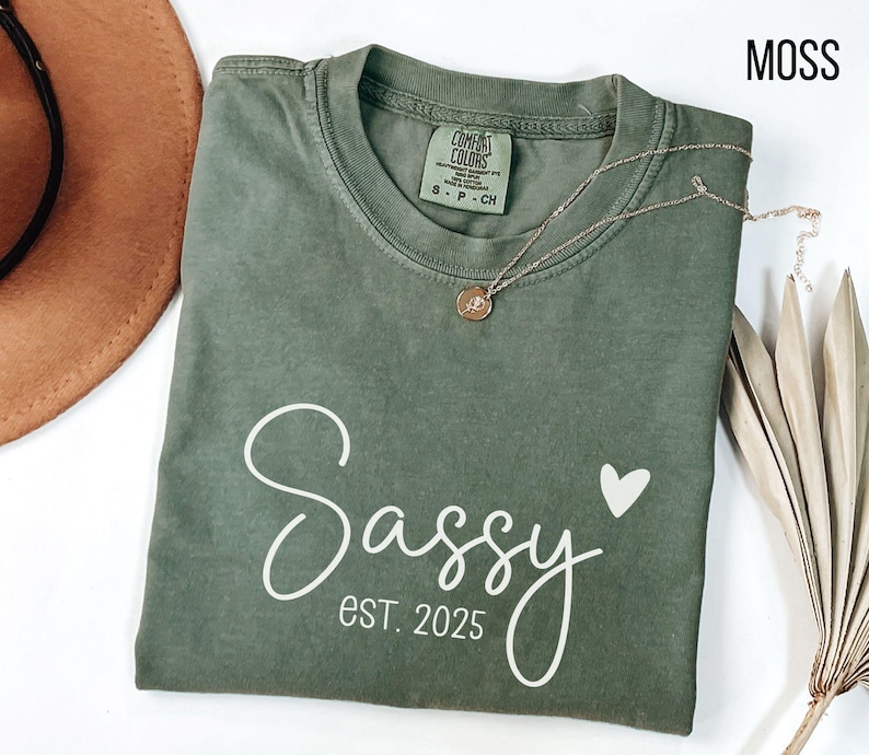 May include: A moss green t-shirt with the text "Sassy est. 2025" and a small white heart printed on the front.