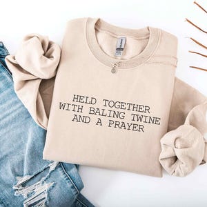 May include: A beige sweatshirt with the text "HELD TOGETHER WITH BALING TWINE AND A PRAYER" in black. The sweatshirt is folded and paired with light blue ripped jeans and a silver necklace.